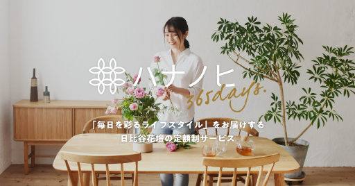 A woman sits at a light wood table with a simple vase containing flowers and a small potted plant