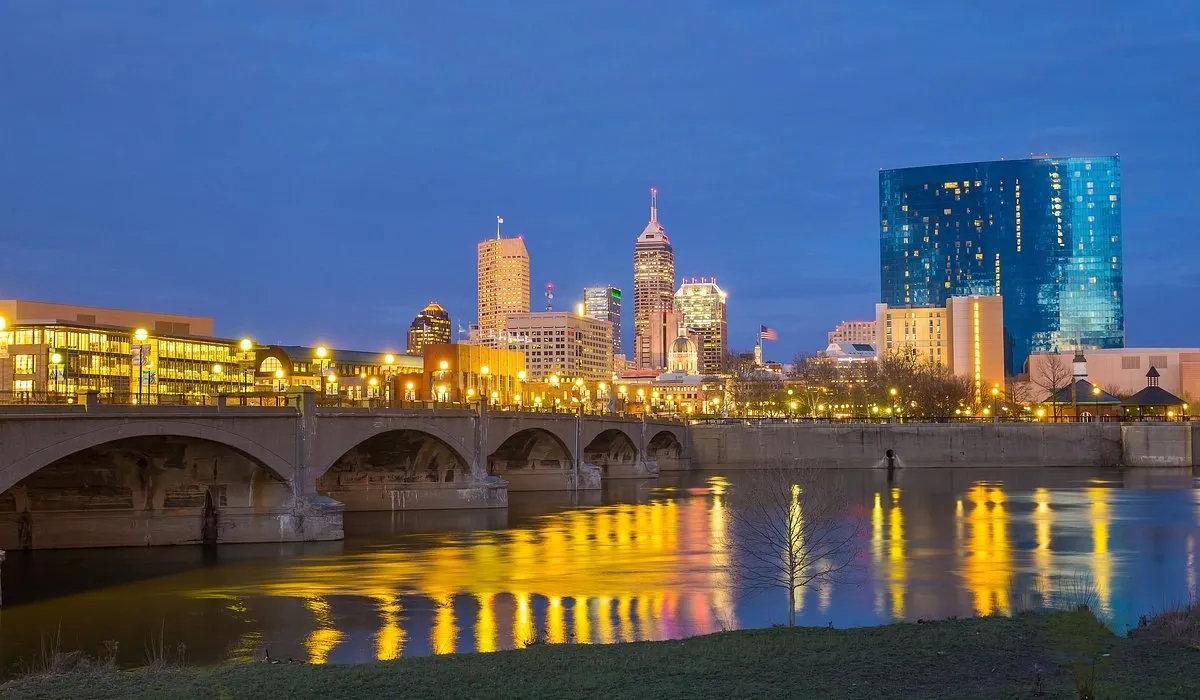 A prominent blue skyscraper reflects in the river alongside an arched bridge overlooking the Indianapolis cityscape at night