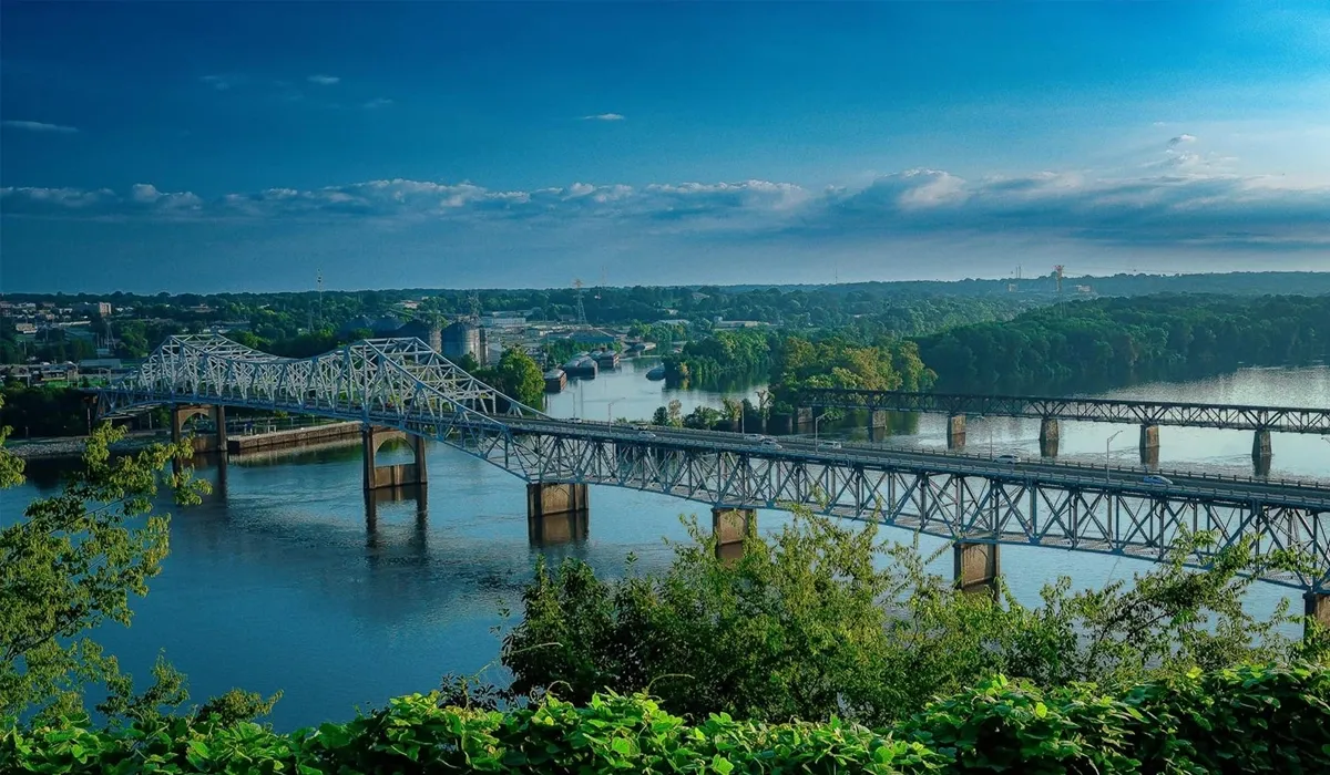A steel bridge spans a wide river reflecting the sky and distant city skyline providing an overview of Muscle Shoals Alabama