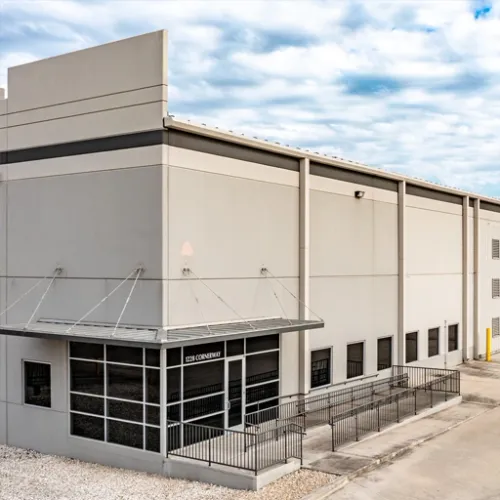 A modern gray warehouse building sits on a paved lot with a black metal fence and minimal landscaping The buildings large