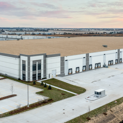 A large industrial warehouse complex with expansive gray buildings sits on a snowy expanse Green lawns and a small security