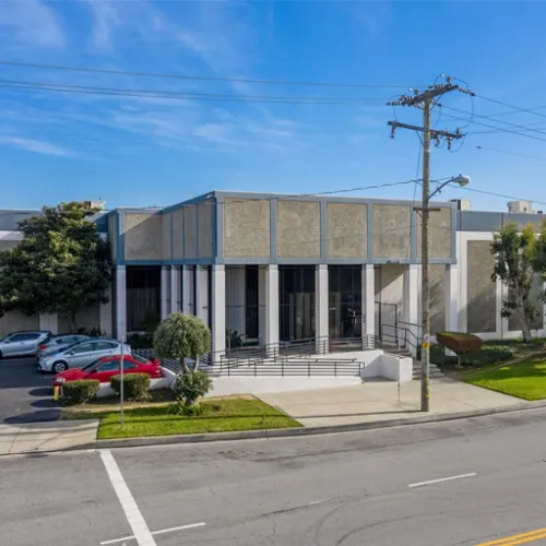 A concrete building with a small lawn and a street sign sits on a city block A white wall and a small tree are visible