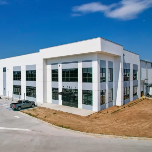 A modern white warehouse building with large windows sits on a paved lot near a dirt shoulder The buildings architecture is