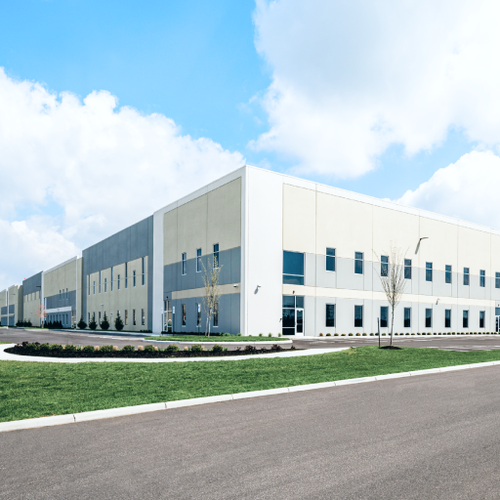 A modern industrial complex featuring large white buildings with blue accents sits on a paved roadway and grassy area